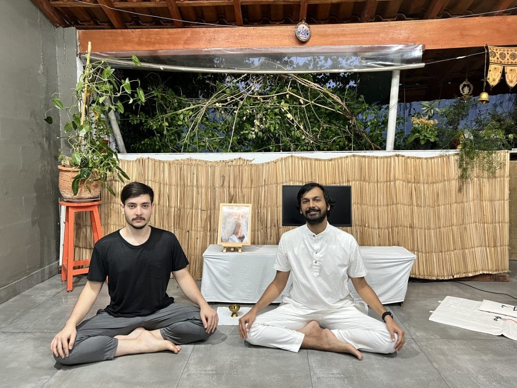 Two men sitting cross-legged on a patio, smiling. one is in black, the other in white; behind them is a makeshift altar with a framed photo.