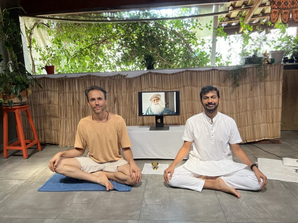 Two men in white clothing sitting cross-legged in a serene indoor yoga studio with a portrait of an elderly man displayed between them.
