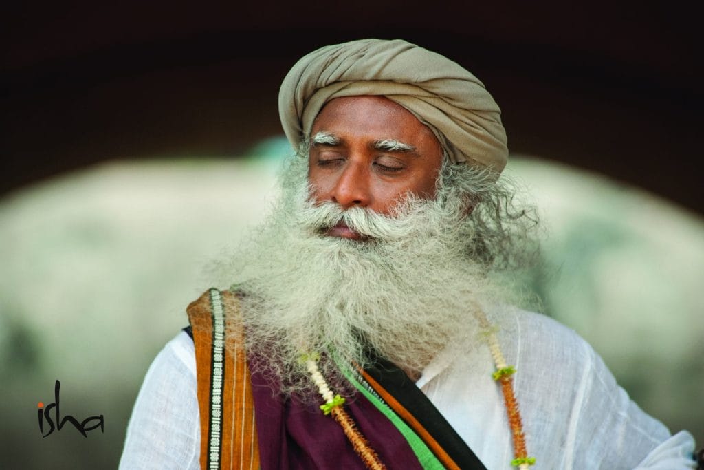 A person with long white hair and beard, wearing a beige turban and colorful clothing, stands outdoors with eyes closed. The word "isha" is visible in the bottom left corner.