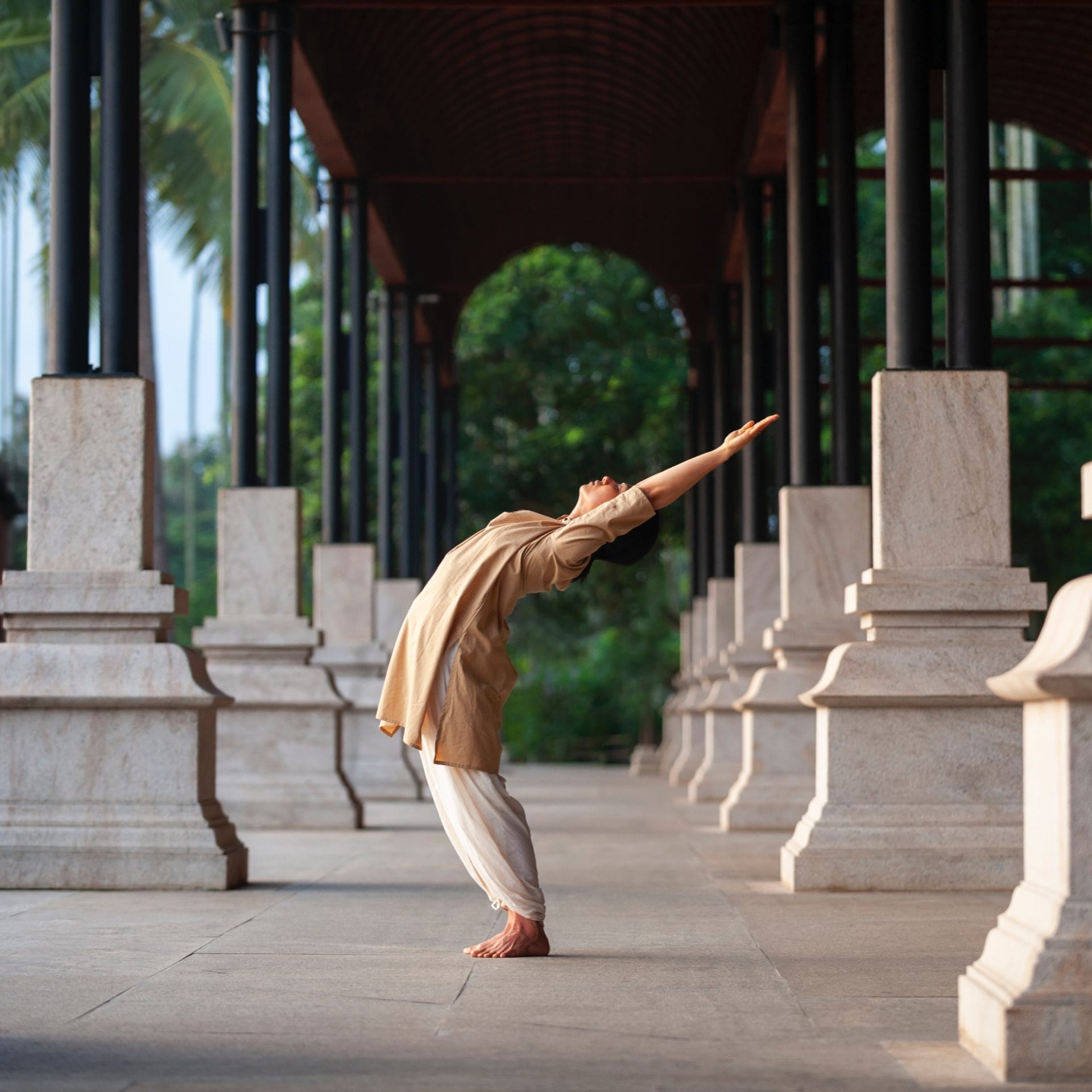 A person stands on a stone walkway between columns, performing a backward bending yoga pose with arms extended overhead.